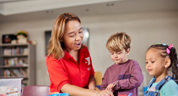 Preschool teacher with a preschool aged boy and girl sitting around a table. The teacher is pointing to the an item on the table,