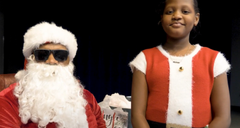 Young girl in Santa shirt next to Santa in sunglasses.