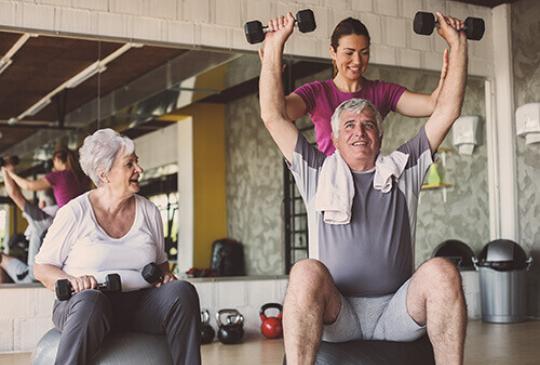 A YMCA trainer helps two active older adults sitting on stability balls lift dumbbells.